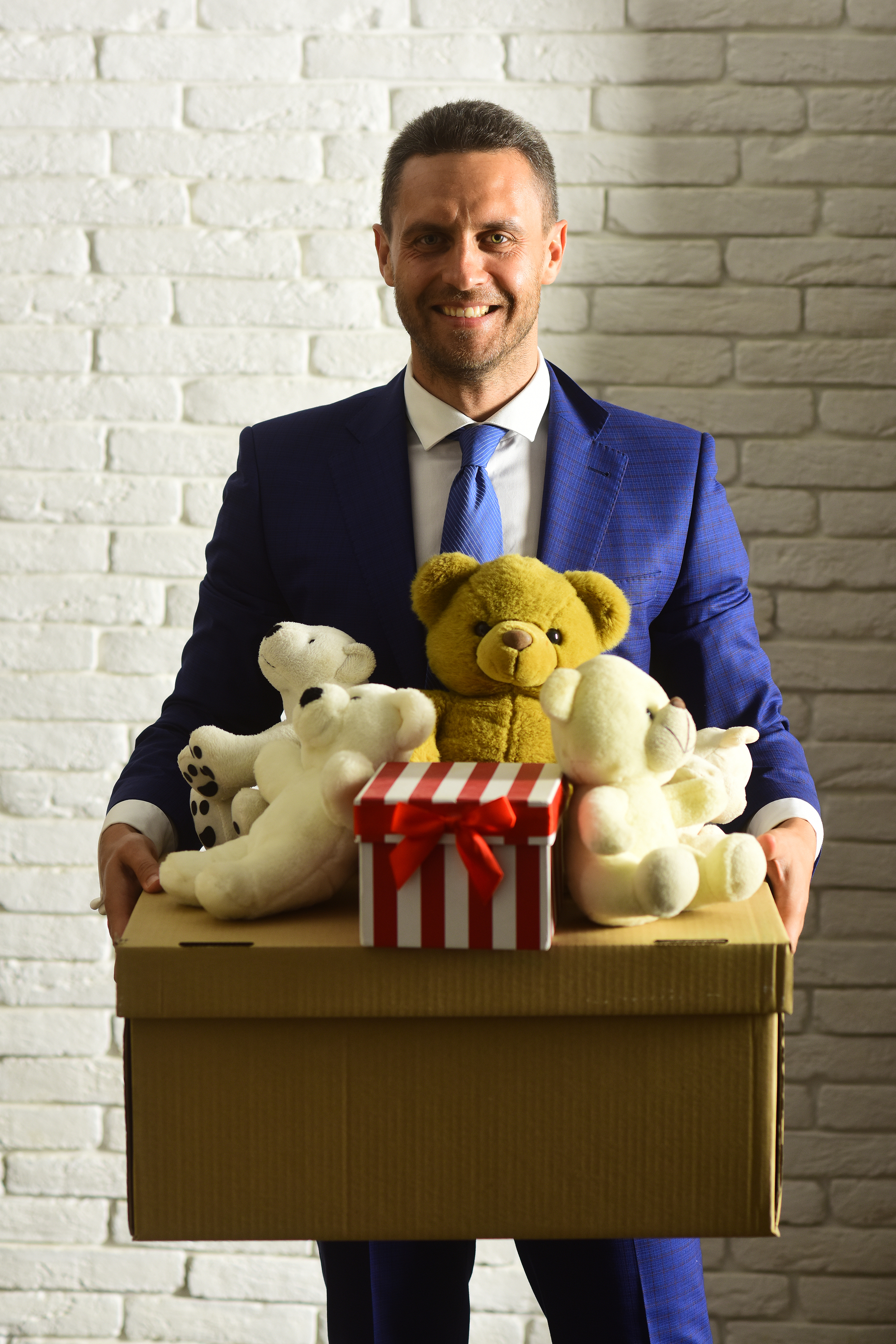 Businessman wears smart suit and tie. Business and volunteering concept. Executive holds box full of toys and Christmas present on white brick wall background. Man with smiling face makes donation.